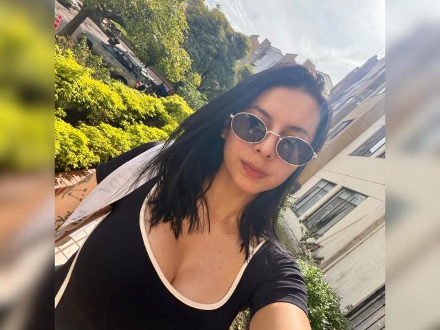 IsabellaJames, con i capelli neri, indossa occhiali da sole rotondi e una maglietta nera con bordi bianchi, mostrando il seno in un selfie scattato all'aperto in una giornata di sole..