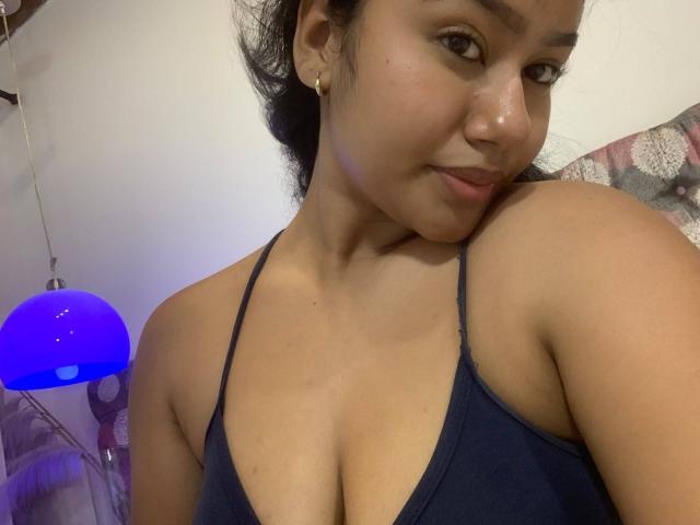 NoiraVale with dark hair, tan skin, and small gold earrings, wearing a navy blue halter top, showing her cleavage and smiling slightly, in a casual selfie.