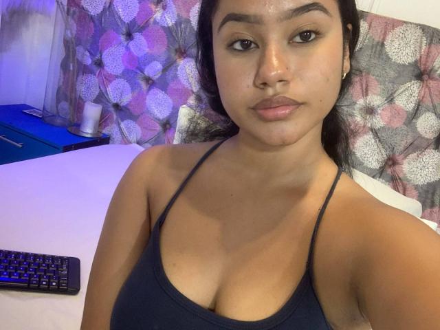 NoiraVale with medium brown skin, dark hair, and small breasts, wearing a black spaghetti-strap top, looking at the camera with a neutral expression. Keyboard on the table.