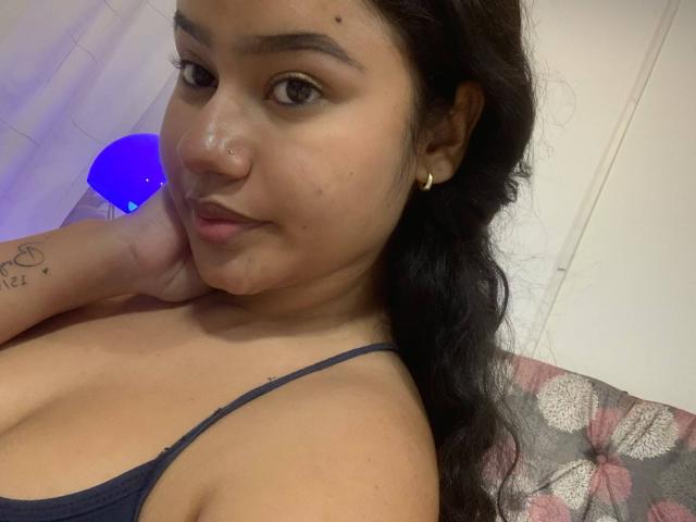 NoiraVale with brown skin, black hair, small gold hoop earrings, blue spaghetti strap top, hand on cheek, subtle smile, small boob tattoo, visible cum on nose.
