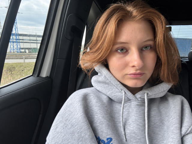 Ardelle Cooksey, com cabelo ruivo, olhos verdes e um moletom cinza claro, sentada em um carro, olhando ligeiramente para o lado com uma expressão neutra. Seus seios são pequenos..