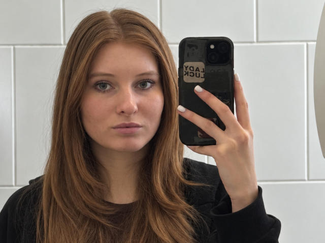 ArdelleCooksey with straight, brown hair, light skin, and blue eyes, holding a black phone with a sticker, wearing a black shirt, looking serious in a mirror selfie.