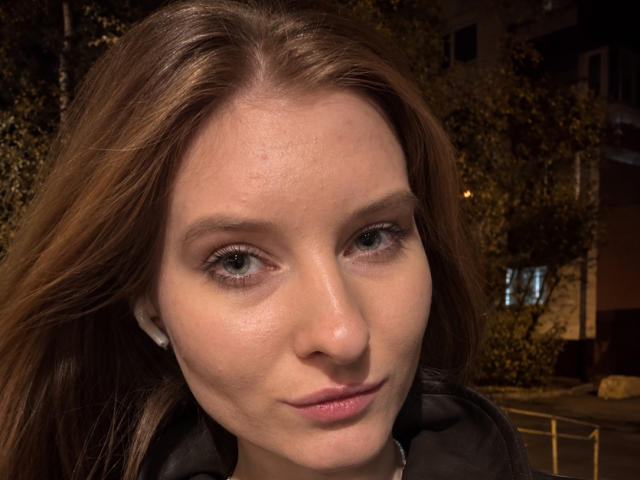 O rosto de Ardelle Cooksey, com cabelo castanho, olhos verdes e pele clara, exibindo um sorriso discreto, vestindo uma jaqueta preta e usando pequenos brincos brancos, em um ambiente noturno..
