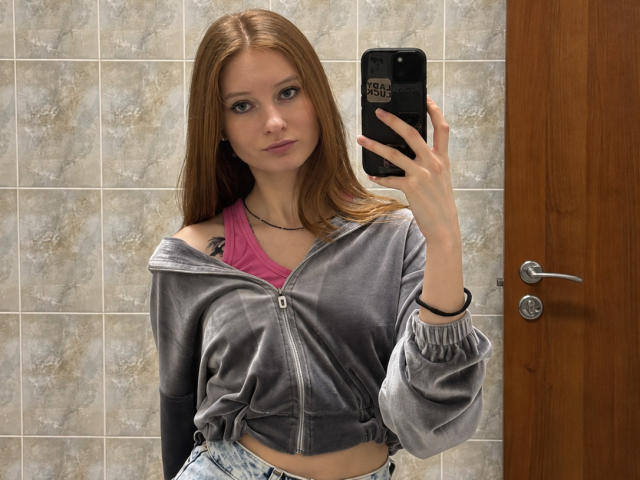 ArdelleCooksey with long brown hair, small tits, pink bra, gray zip-up hoodie, holding phone in mirror, wood door, tiled wall, looking at camera, slight smirk.
