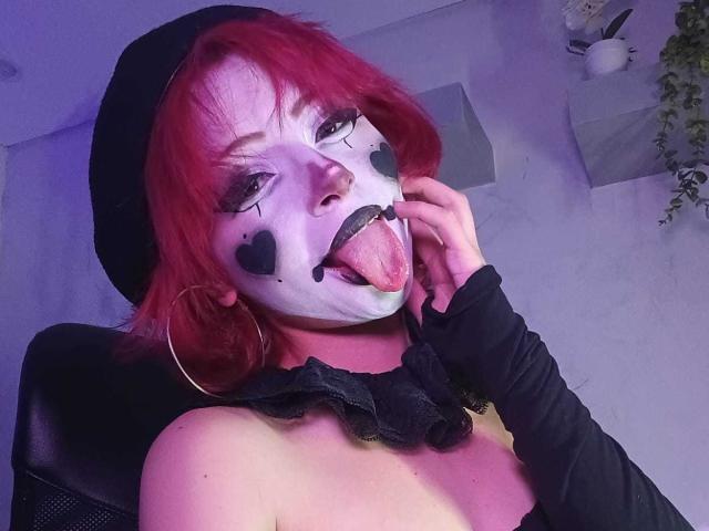 ViolettClay with red hair, black face paint, and heart shapes on cheeks, sticking out tongue, wearing black top, ruffled collar, and black beret, in a chair.