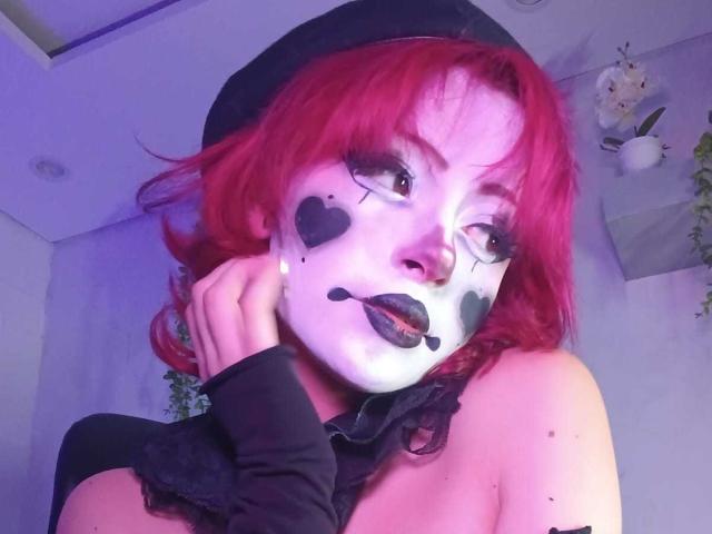 ViolettClay avec des cheveux roux, un eye-liner noir, du maquillage en forme de cœur, un masque blanc, du rouge à lèvres noir, un manchon noir, un voile noir, une main sur la joue, un éclairage bleu-rose, des fleurs en arrière-plan..