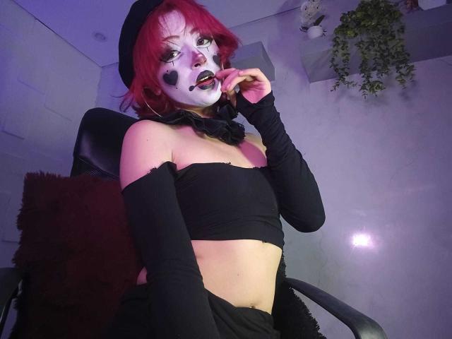 ViolettClay, com cabelo ruivo, maquiagem branca no rosto, batom preto e piercing no nariz, vestindo uma blusa preta, mangas compridas e um chapéu, tocando os lábios, sentada em uma cadeira..