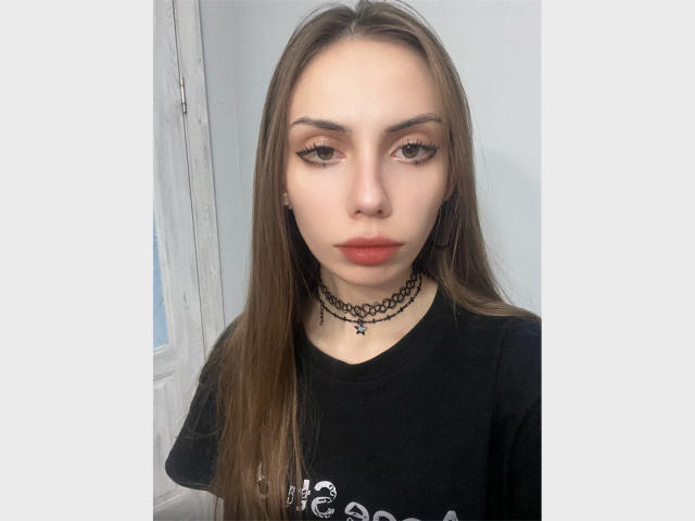 JudyPalas, avec ses longs cheveux bruns, son collier noir et sa chemise noire, a de petites seins, des lèvres rouges pulpeuses et des yeux marrons. Elle regarde directement l'appareil photo avec une expression neutre..