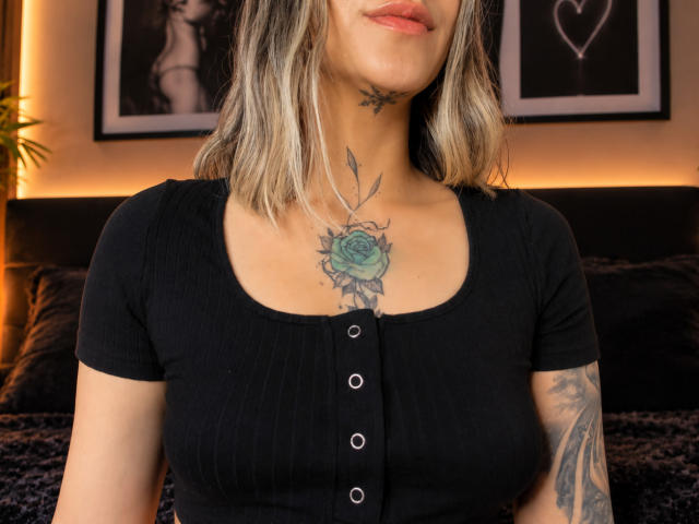 KathyM, met blond haar dat tot op haar schouders reikt, een zwart gestreept shirt met knopen, waarbij een groene roos tatoeage op haar borst zichtbaar is, en een tatoeage op haar linkerarm, in een slaapkamer..