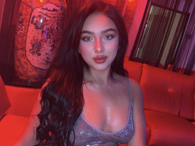 LilyConorr with long dark hair, wearing a sparkly grey tank top, showing her shiny boobs, under red and purple lighting, looking directly at the camera.