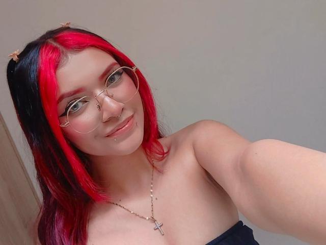 AriaRosel with red and black hair, glasses, nose ring, lip piercing, cross necklace, smiling, bare shoulders, taking a selfie, showing a cute face and cute tits.