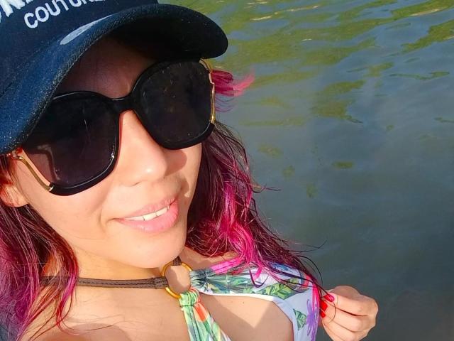 LunaTomson met roze haar, een zwarte pet en oversized zonnebril, lacht naar de camera. Ze houdt een kleurrijke bikini-top vast, waardoor haar kleine borsten zichtbaar zijn. Op de achtergrond is water te zien..