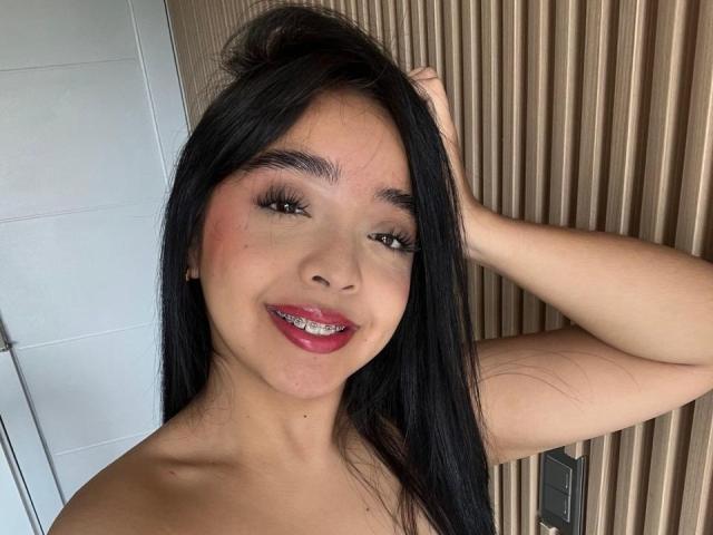 VictoriaKross a de longs cheveux noirs, des yeux marrons, du rouge à lèvres, et elle sourit, une main dans ses cheveux. Elle porte des appareils orthodontiques et a un visage mignon et attrayant, avec un léger rougissement..
