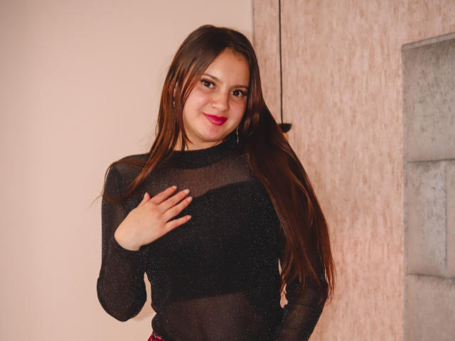 BeccaSimons with long brown hair, wearing a sheer black top, hand on chest, smiling with pink lipstick, standing in front of a textured wall, showing her small tits.