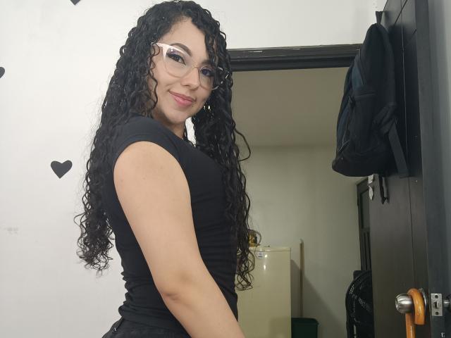 ChloeMonet with curly black hair, glasses, and a black sleeveless top, smiling at the camera, showing her ass, standing near a door with a black bag hanging.