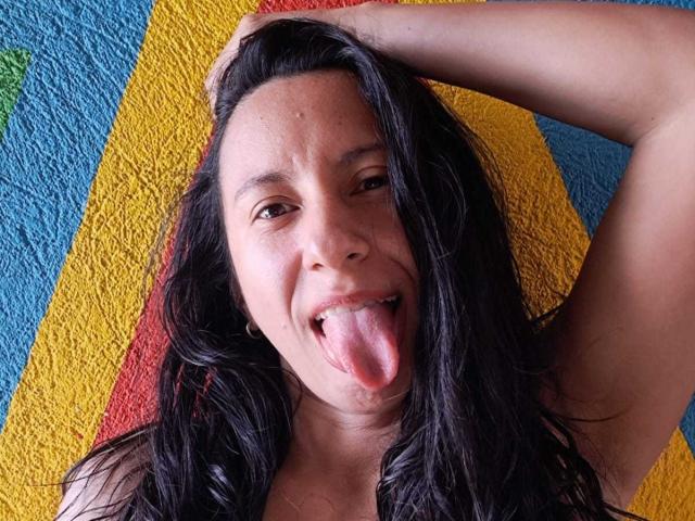 Jade Foster, con lunghi capelli neri, occhi marroni e un sorriso carino, che mostra la lingua, alza il braccio destro, su un asciugamano colorato..