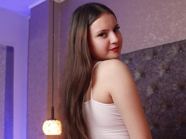 BeccaSimons with long brown hair, white tank top, looking over shoulder, smiling, showing her ass, in a bedroom with a tufted headboard and a hanging lamp.