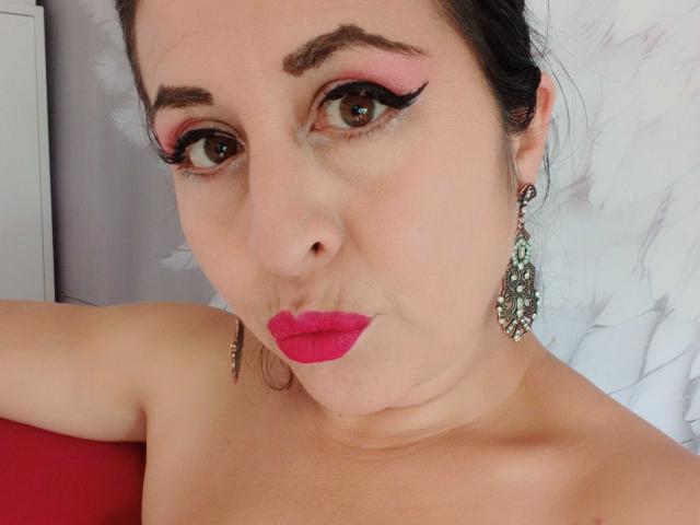 ScarletMilfX hat rosigen Lidschatten, schwarzen Eyeliner, roten Lippenstift und trägt Diamant-Ohrringe. Sie zieht die Nase leicht hoch und macht eine Schmollmund. Ihre Brüste sind außerhalb des Bildausschnitts, und sie ist oben ohne..