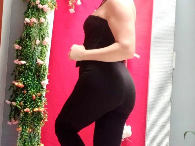 ScarletMilfX lleva un top negro sin tirantes y pantalones negros ajustados, mostrando su trasero y sus pequeños senos firmes, con una guirnalda floral verde en el lado izquierdo..
