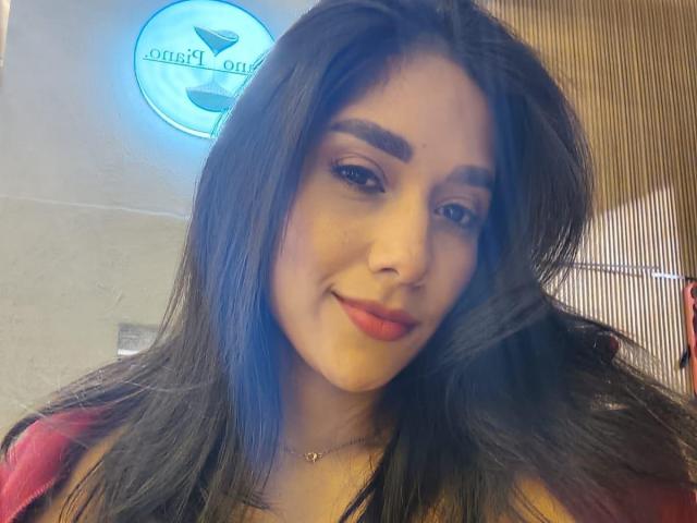 BrianaHarpers a de longs cheveux noirs, des sourcils épais, du rouge à lèvres et un léger sourire. Elle porte un haut rouge avec un collier en or. Ses seins sont partiellement visibles..