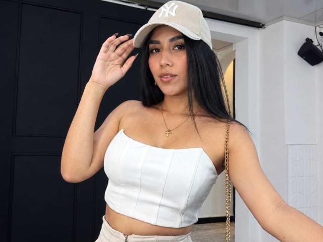BrianaHarpers, avec de longs cheveux noirs, portant un haut blanc sans bretelles, une casquette beige avec le logo NY, un collier en or et un pantalon beige, tenant sa casquette, montrant ses seins..