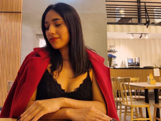 BrianaHarpers com seios médios, sutiã de renda preta, casaco vermelho, cabelo preto, batom vermelho, braços cruzados, sentada em um café, cadeiras de madeira, sorriso suave, relaxada, luz quente..