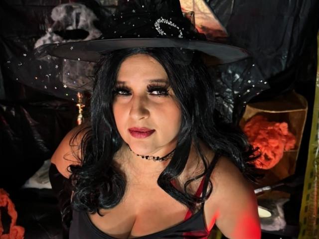 KerlyDolly con i capelli neri e ondulati, che indossa un cappello da strega, una collana nera e rossetto rosso, mostra il suo seno prosperoso in una maglietta nera, con un'espressione seria e leggermente compiaciuta..