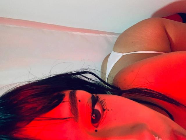 HannaThorne with dark hair, red light, and white thong, lying on bed, showing her big ass and large tits, black eyebrow tattoo, and nose ring.