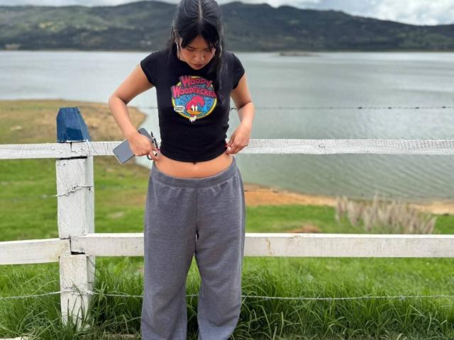 HannaThorne with dark hair, black shirt, grey pants, lifting shirt to show belly, standing in front of white fence, holding small device, near green grass and water.