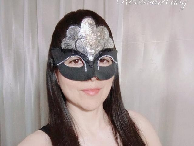 RossanaHairy with black mask, silver sequin flower on forehead, brown eyes, black straight hair, pale skin, pink lips, wearing black sleeveless top, soft smile, white curtain background.