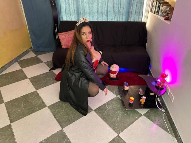 LexyDom kneels on checkered floor, wearing black leather robe, red lingerie, black stockings, and tiara, with large tits, long brown hair, and red lipstick, in a Halloween-themed room.