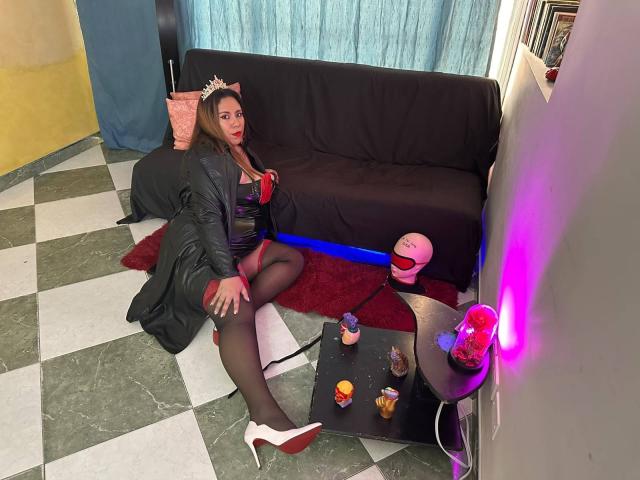 LexyDom with brown hair, wearing a black leather dress, red bra, black stockings, white heels, crown, sitting on floor, black couch, red rug, neon pink light, and drinks.