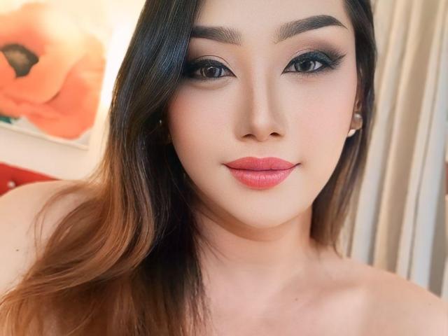 LexyGrayCum with smooth, fair skin, dark brown hair, and pink lipstick, has sultry, black-lined eyes, and small, pearl earrings. Boobs not visible. Face-centered, smiling slightly.