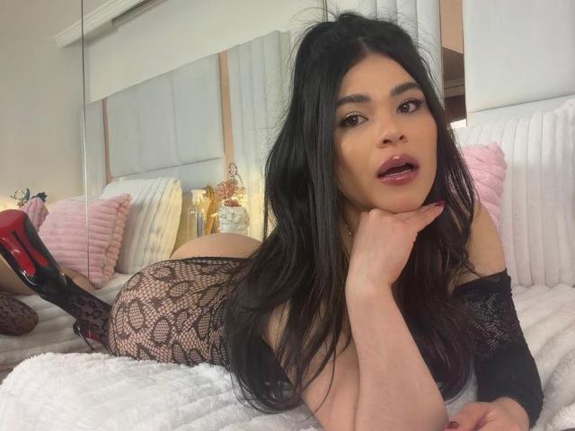 PaulinaValley, met lang zwart haar, liggend op een bed, gekleed in kant lingerie en zwarte kousen, haar hand onder haar kin, rode zolen, kijkend naar de camera, met licht open lippen..