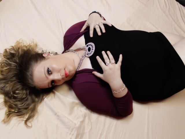 VanessaSquirtt lies on a bed, wearing a black dress with purple sleeves, hands on her chest, necklace, and ring, blonde hair splayed, red lipstick, looking at camera.