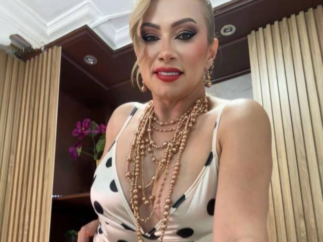 MisseScarlet, avec ses cheveux blonds, son rouge à lèvres rouge et sa robe à pois, portant plusieurs colliers de perles, souriante, montrant ses seins, dans une pièce avec des rideaux beige et des fleurs violettes..