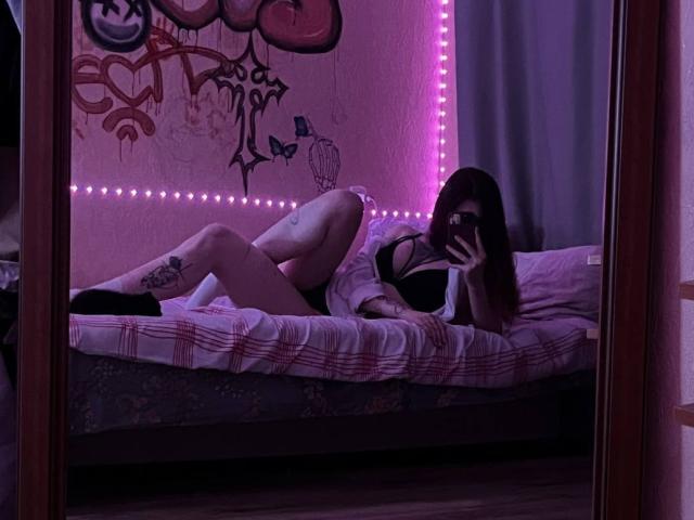 MandiSalais with pale skin, long brown hair, black lingerie, tattoo on left thigh, lying on bed, taking mirror selfie, purple LED strip, bed with pink and white sheets.