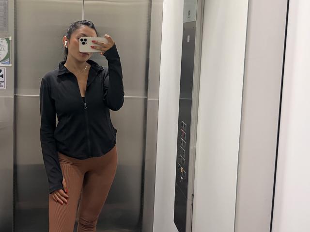 AmeliRoux in an elevator, wearing a black zip-up shirt and brown leggings, taking a mirror selfie with her phone, covering her face. She's got small tits and a cute butt.