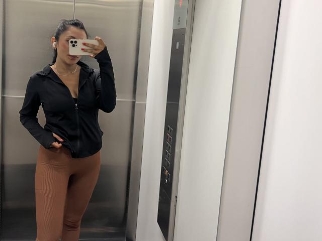 AmeliRoux dans un ascenseur, portant un haut noir à fermeture éclair, un legging marron taille haute, une main dans la poche, tenant un téléphone, se prend en photo devant un miroir, cheveux longs et foncés attachés en queue de cheval, maquillage discret, portant un collier..