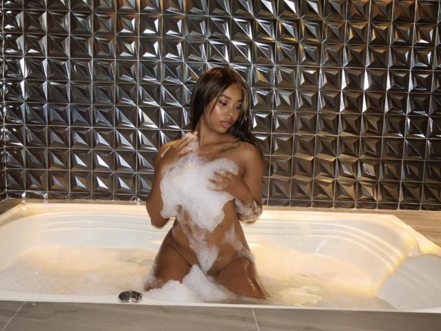 EmmaLatin, nude, sitting in a bubble-filled bathtub, covering her tits with soap suds, dark brown hair, tattoo on right side, looking down, wet skin, attractive, sexy, seductive.