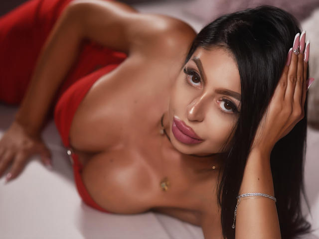 AvaNoah, con i capelli scuri, sdraiata, indossa un vestito rosso, che mette in mostra il suo seno prosperoso, con una mano tra i capelli, rossetto rosa e un braccialetto d'argento, mentre guarda verso la fotocamera..