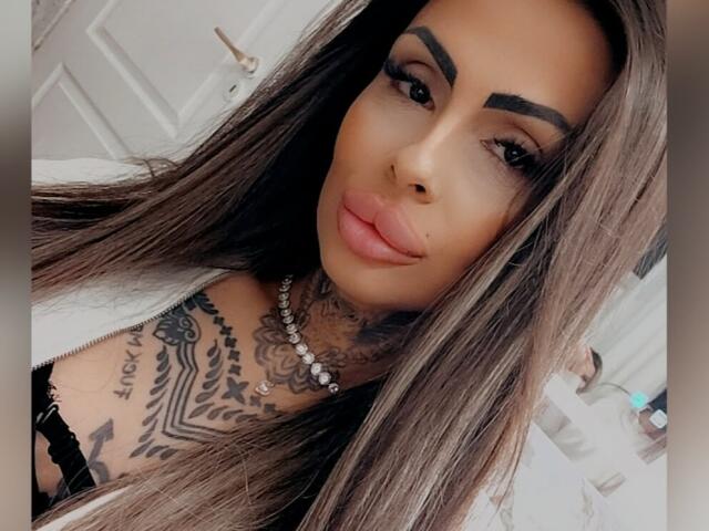 DenyChantal with long brown hair, thick eyebrows, and full pink lips. She's got tattoos on her neck and chest, wearing a black lace top and a pearl necklace.