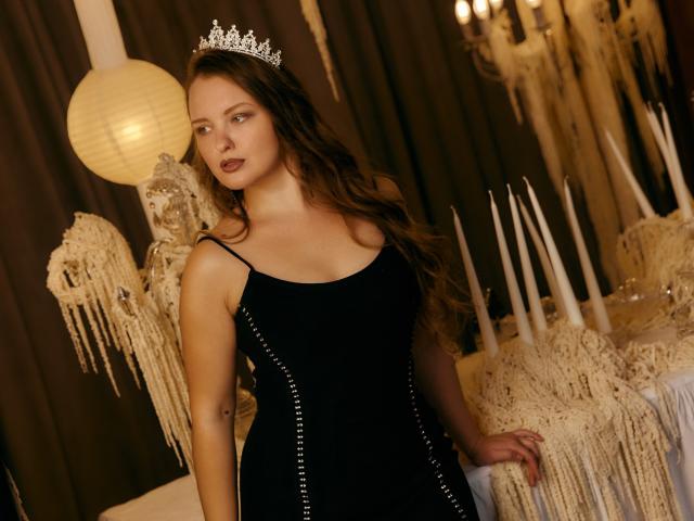 AliceSagkew with long brown hair, wearing a black dress with silver studs, a silver tiara, standing in front of white candles and hanging paper decorations, looking serious.
