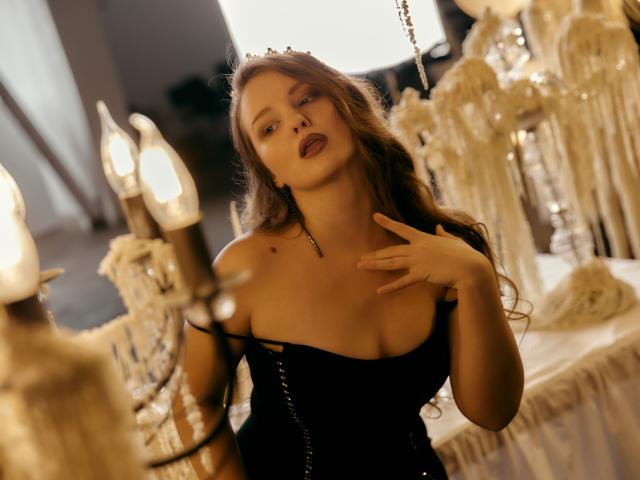 AliceSagkew with wavy brown hair, wearing a black strapless dress, touching her chest, showing her tits, in a room with candles and white fabric. Soft, seductive expression.