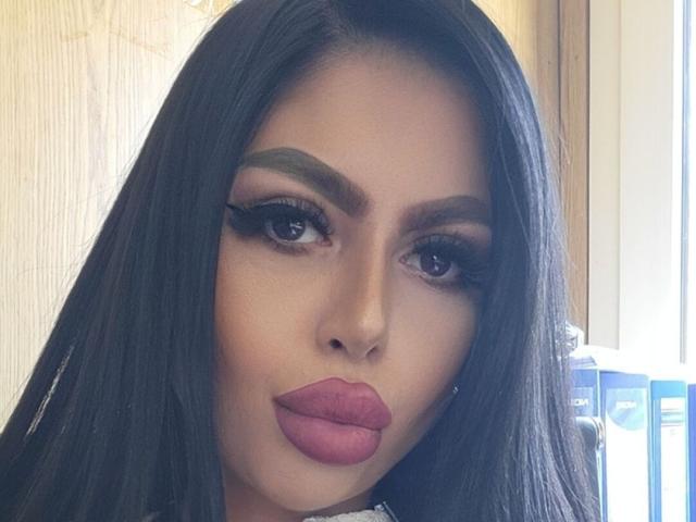 AvaNoah tiene el pelo largo y negro, cejas gruesas, delineador de ojos oscuro y lápiz labial rosa. Sus ojos son oscuros y está mirando ligeramente hacia un lado, con un ligero puchero..