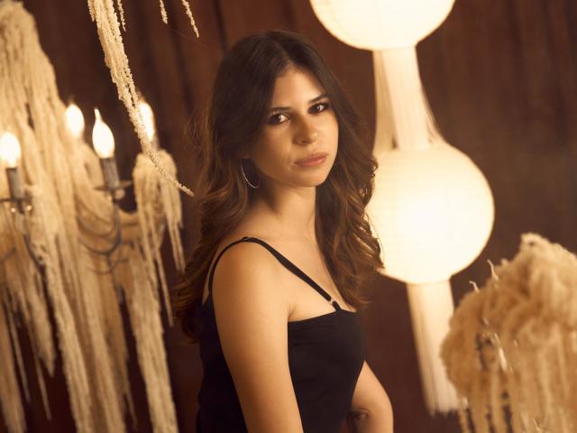 CaseyTerorib with wavy brown hair, wearing a black spaghetti strap top, standing in a dimly lit room with warm lighting and hanging decorations. She has small tits and a tattoo.