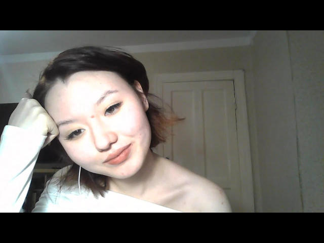 EmaPepper with light skin and brown hair, smiling slightly, wearing a white off-shoulder shirt, head resting on hand, small nose, light makeup, and thin earrings.