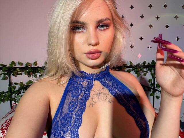 BaeonLive with blonde hair, blue lace halter top, exposing tits, holding pink nail file, tattoo above chest, pink lips, looking at camera, in a bedroom.