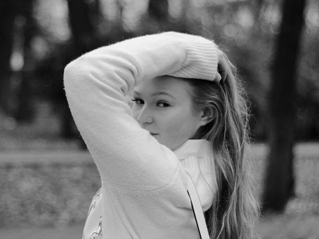 Black-and-white photo of AkilahGhil with long hair, wearing a sweater, hand on head, looking sideways. Her tits are small, and her expression is slightly serious.