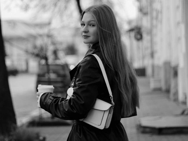 Black-and-white photo of AkilahGhil with long, straight hair, holding a coffee cup, wearing a jacket, and a small crossbody bag, looking over her shoulder with a slight smile.